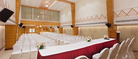 Un salón de conferencias con filas de sillas blancas, una larga mesa con mantel rojo y una gran lámpara de araña.