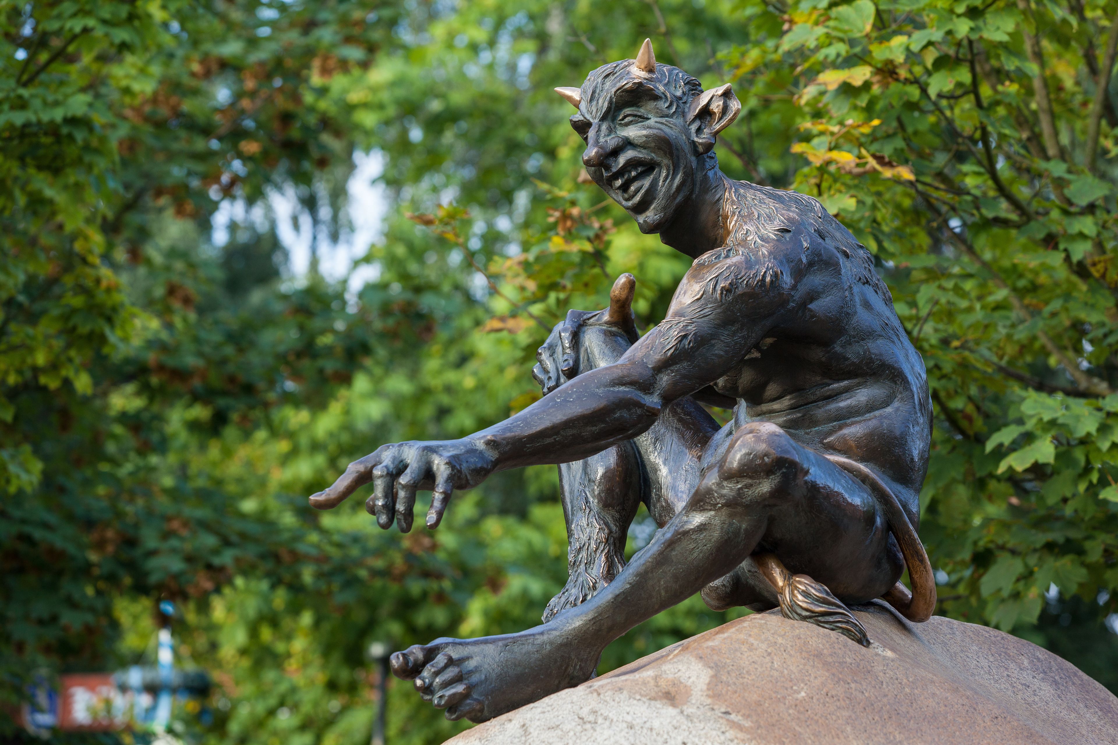 Een bronzen beeld van een zittend figuur met uitgestrekte armen, voor een achtergrond van groen gebladerte.