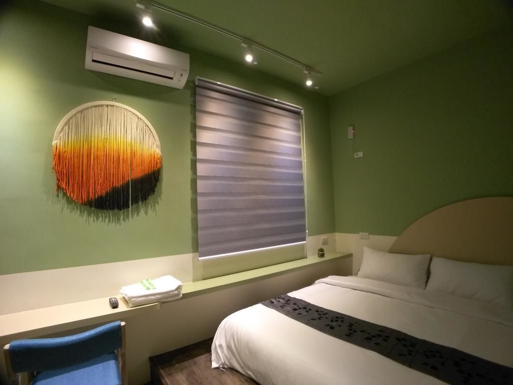 A hotel room with a bed, a desk, a chair, a wall-mounted air conditioner, a window with blinds, and a wall art piece.