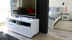 A modern bedroom with a TV on a white stand, a bed with grey bedding, and a small bedside table with a lamp. - Baltazar Studio Apartment (Zagreb)