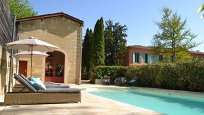 A pool area with lounge chairs and umbrellas, a stone building with a red door, and a clear blue sky. - Château Pont Saint-Martin (Leognan)