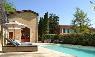 Un área de piscina con sillones y sombrillas, un edificio de piedra con una puerta roja y un cielo azul despejado.
