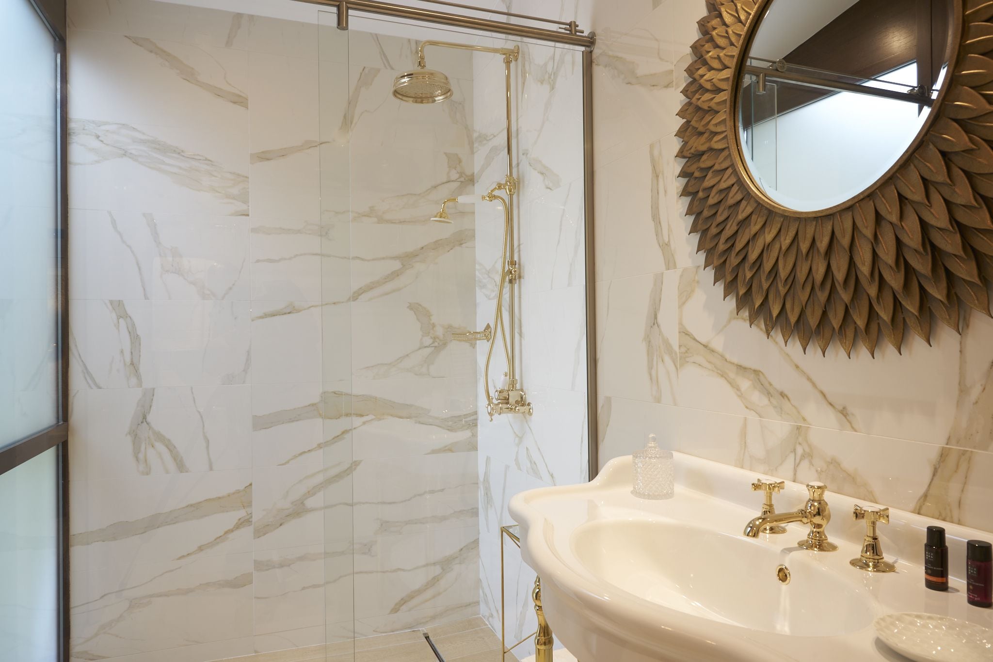 Une salle de bain avec un mur en marbre, une douche dorée, un lavabo avec des robinets dorés et un miroir décoratif.