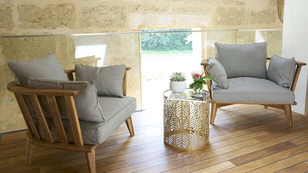 A room with two wooden chairs with grey cushions, a glass coffee table with a decorative metal base, and a stone wall. - Château Pont Saint-Martin (Leognan)