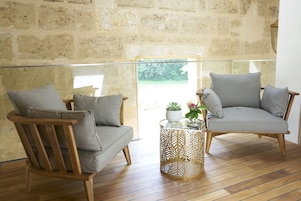 Una habitación con dos sillas de madera con cojines grises, una mesa de centro de vidrio con base de metal decorativo y una pared de piedra.