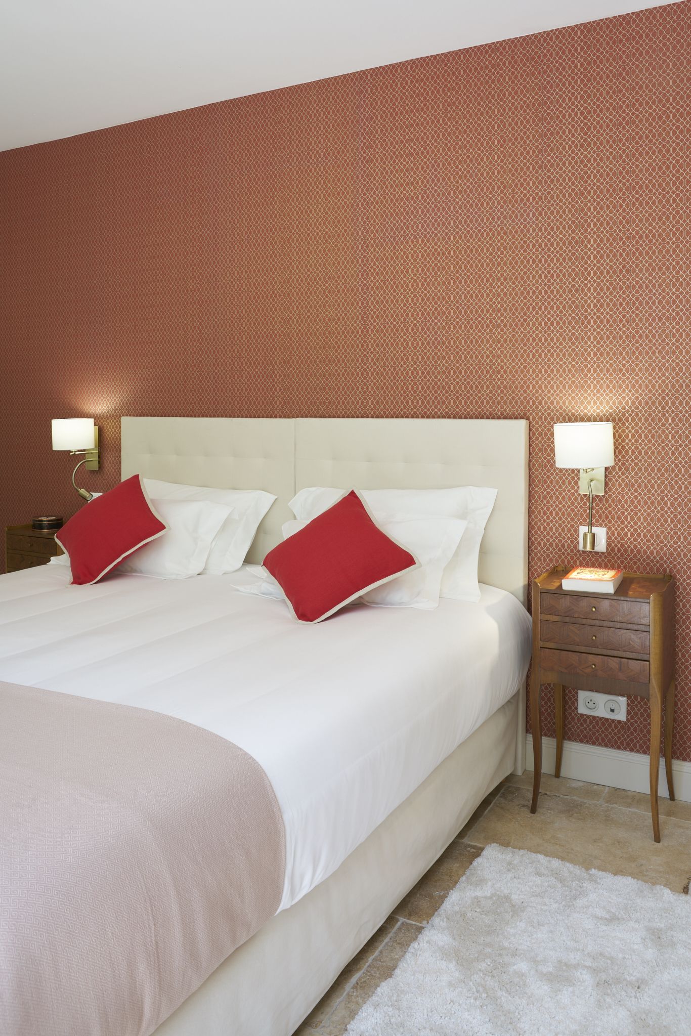 Un lit bien fait, avec des oreillers blancs et rouges, une table de chevet en bois surmontée d'une lampe, et un mur orné d'un motif.