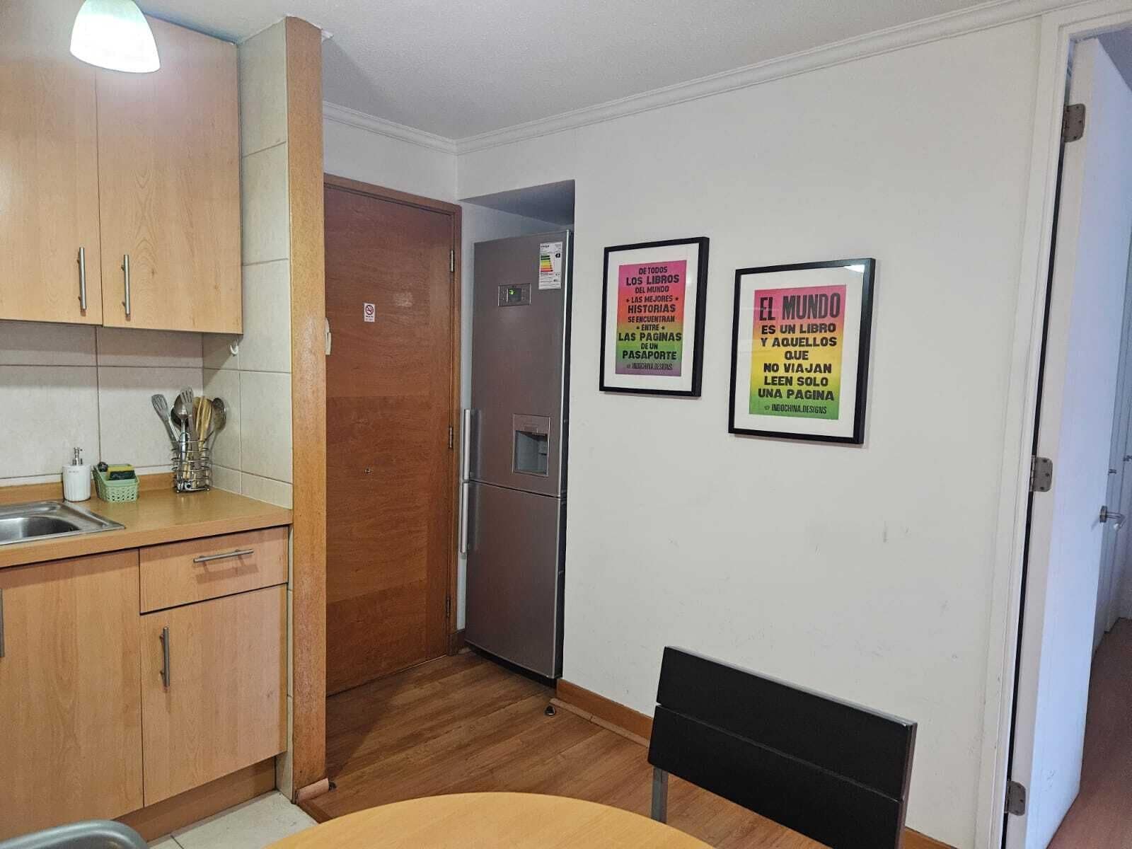 A compact kitchen with wooden cabinets, a stainless steel sink, and a refrigerator. There are two framed posters on the wall.