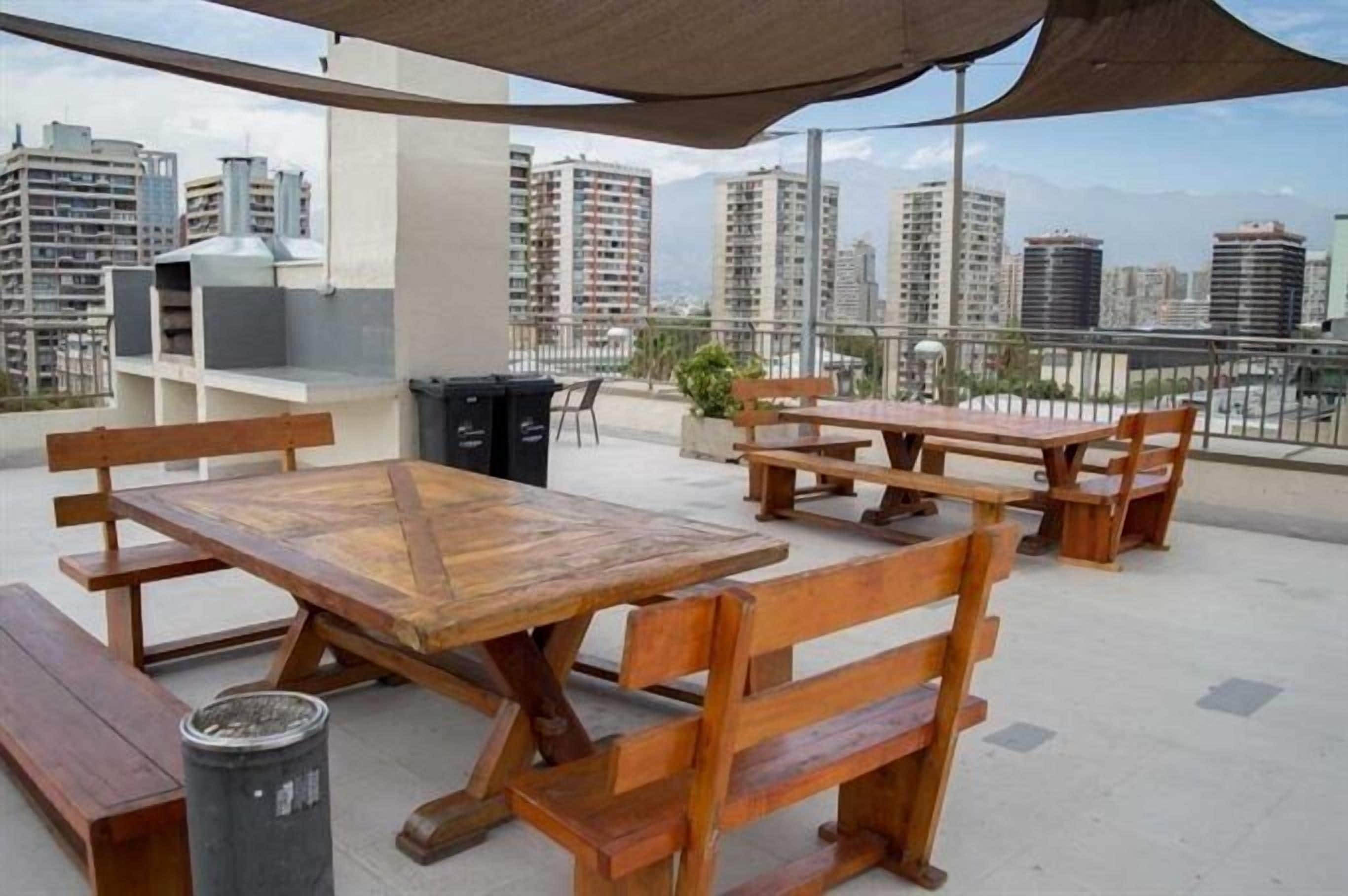 Un patio al aire libre con mesas y bancos de madera, un bote de basura y vistas de edificios de la ciudad.