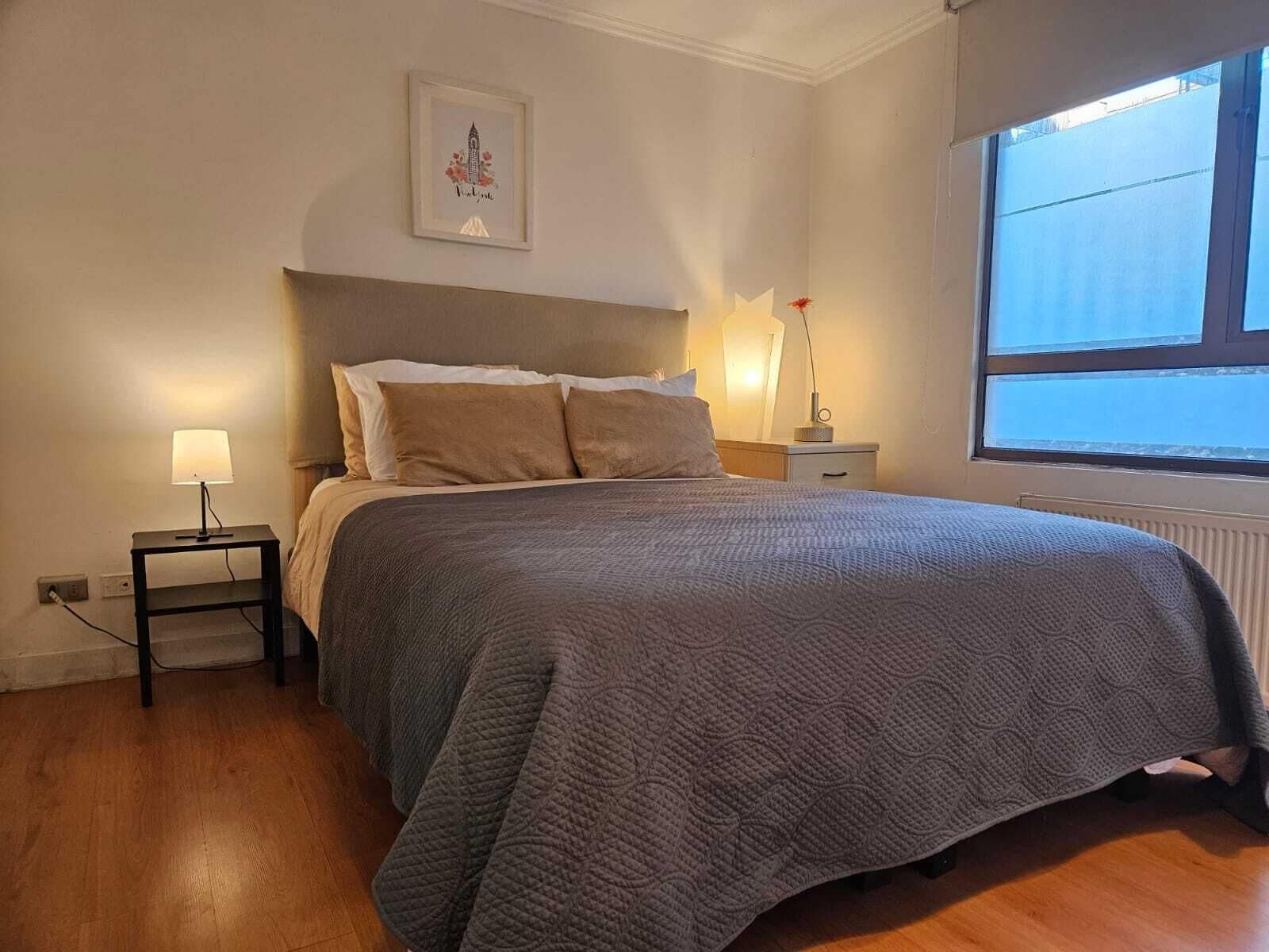 A neatly made bed with a grey quilt, a bedside lamp, a nightstand, and a framed picture on the wall.