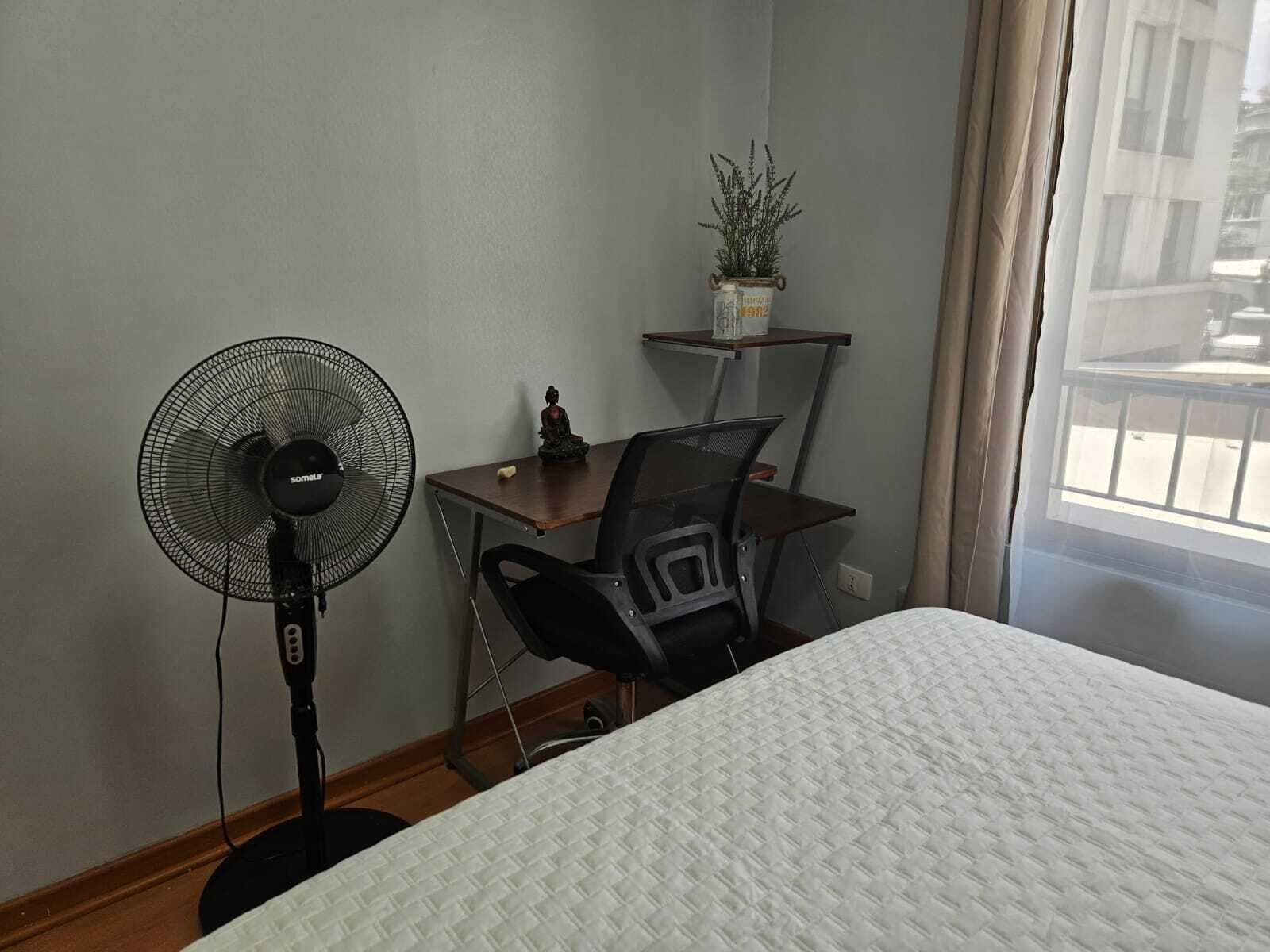 A bedroom with a bed, a desk with a chair, a standing fan, and a window with curtains.