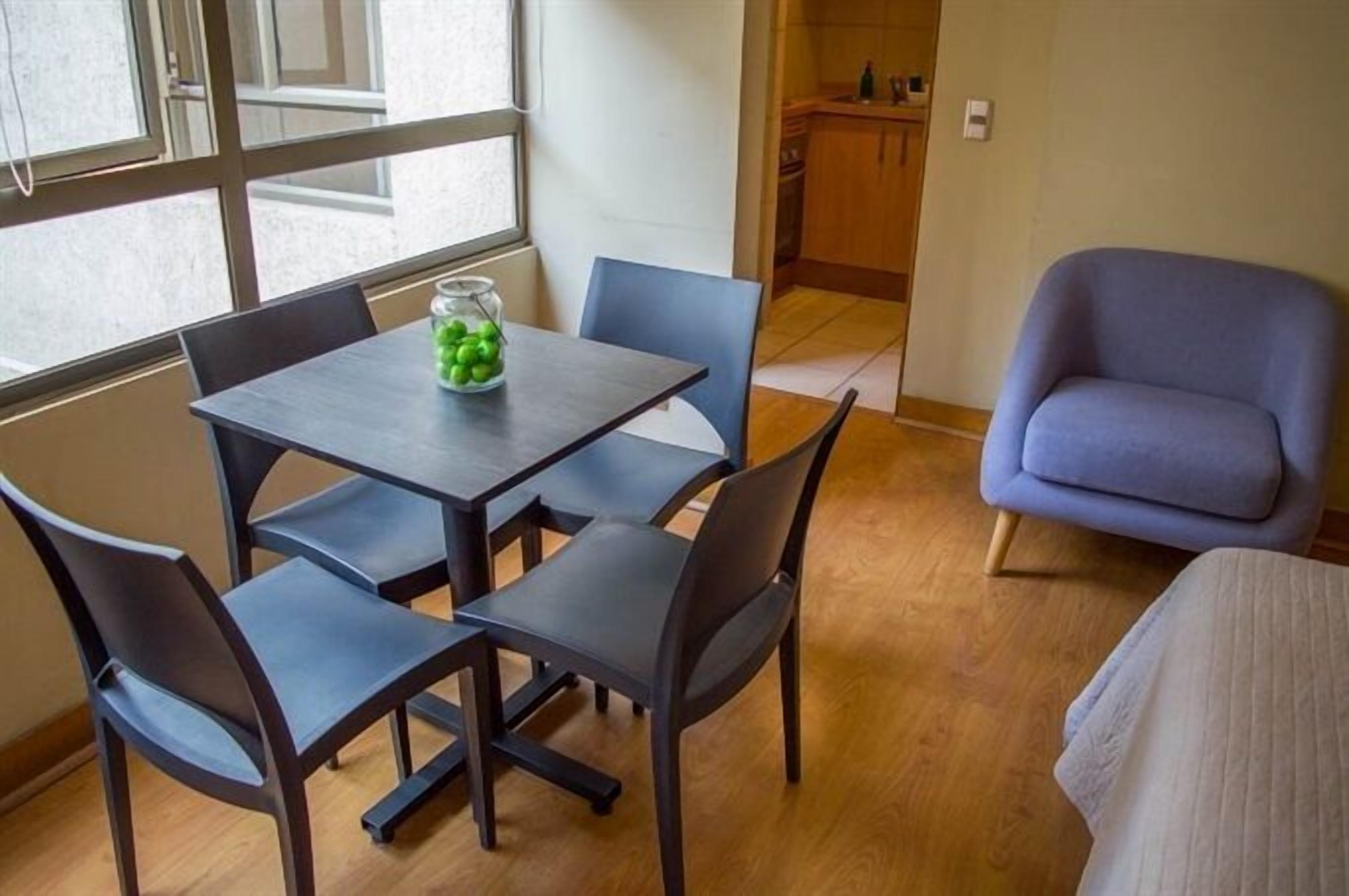 A small room with a table and four chairs, a window, a kitchenette, and a blue armchair.