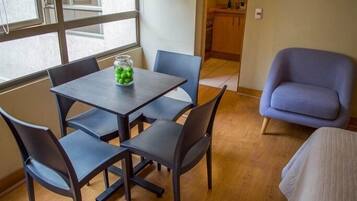 A small room with a table and four chairs, a window, a kitchenette, and a blue armchair.