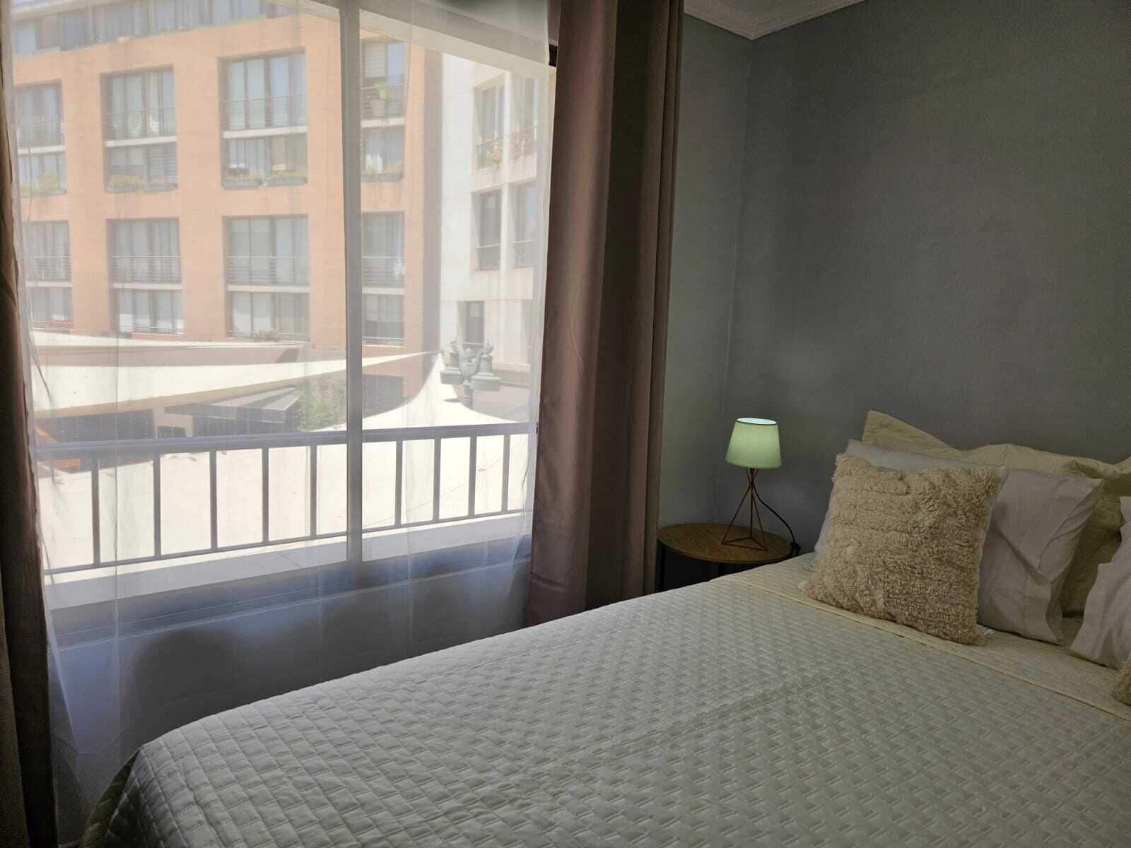 A hotel room with a large bed, a nightstand with a lamp, and a view of a building through the window.