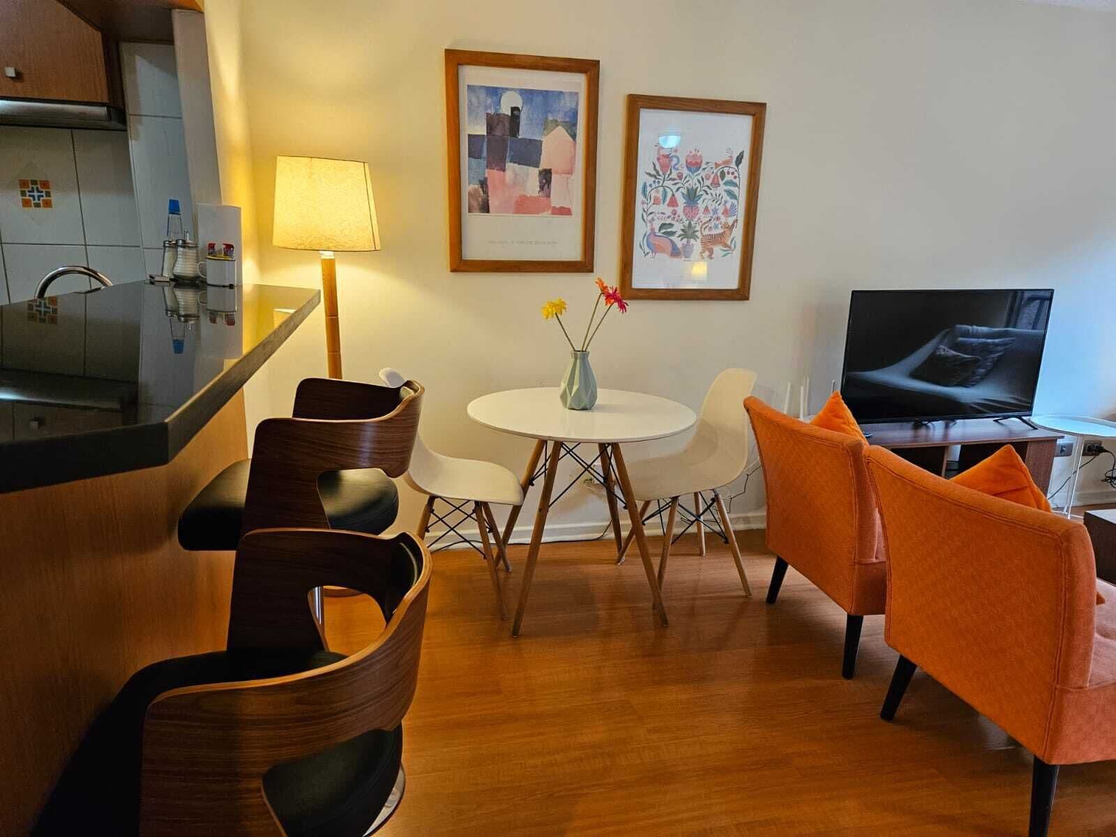 Una sala de estar moderna con zona de cocina, una mesa redonda de comedor con sillas, un sofá y un televisor.