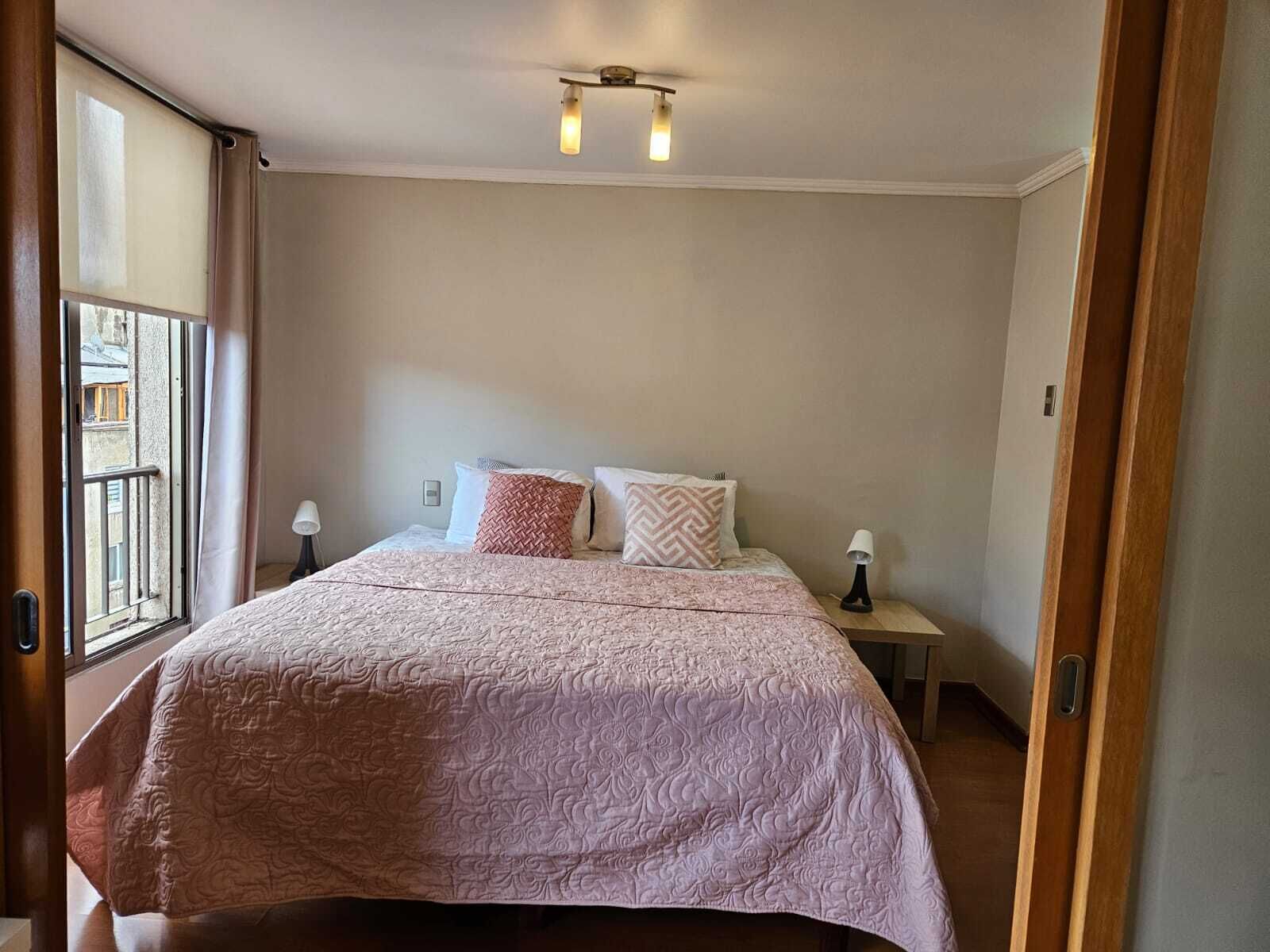A bedroom with a bed, two bedside lamps, a ceiling light fixture, and a window with a view.