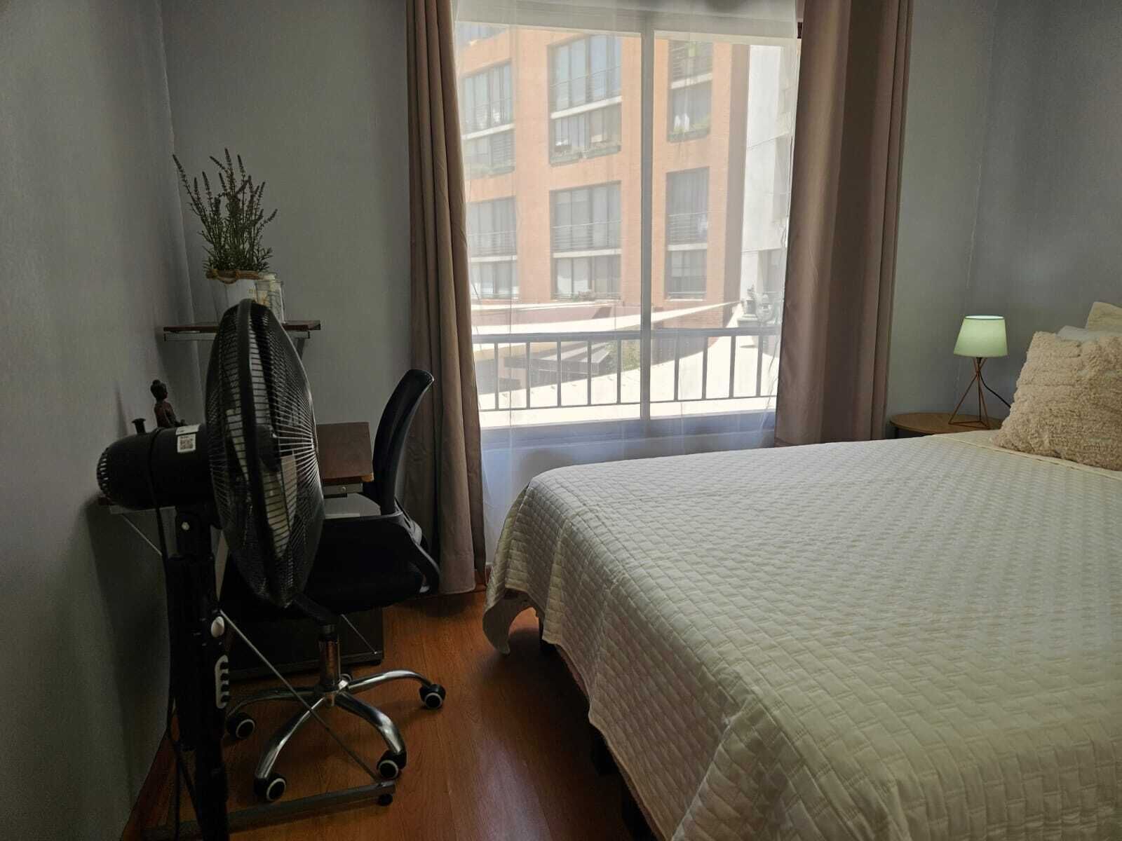 A hotel room with a bed, a desk with a chair, a standing fan, a small plant, a window with curtains, and a view of buildings outside.