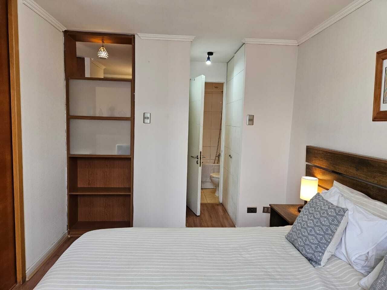 Un dormitorio con cama, mesita de noche, lámpara, armario de madera y baño visible a través de una puerta abierta.