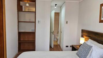 Un dormitorio con cama, mesita de noche, lámpara, armario de madera y baño visible a través de una puerta abierta.