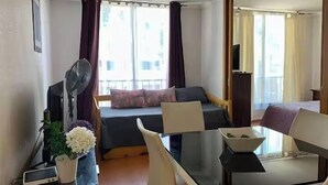 Een hotelkamer met een bed, een bureau met een computer, een stoel, een tafel, een ventilator, een televisie en een raam met gordijnen.