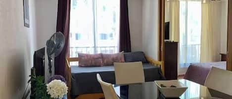 A hotel room with a bed, a desk with a computer, a chair, a table, a fan, a TV, and a window with curtains.