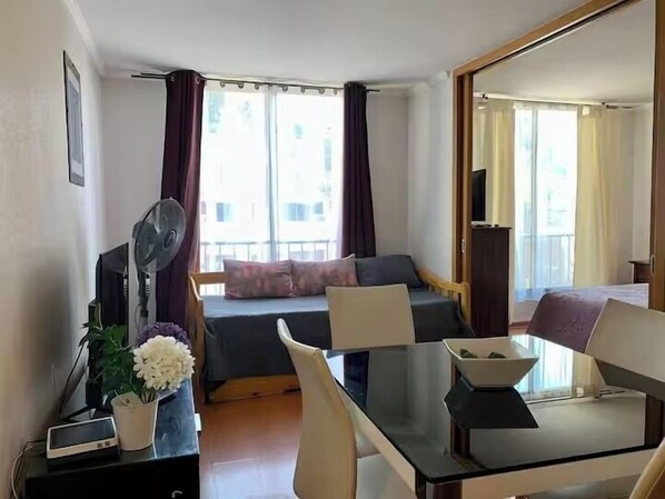 A hotel room with a bed, a desk with a computer, a chair, a table, a fan, a TV, and a window with curtains.