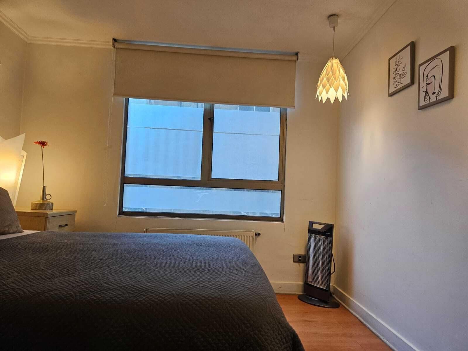 A bedroom with a large window, a radiator, a standing heater, a bed, a nightstand, and a lamp.