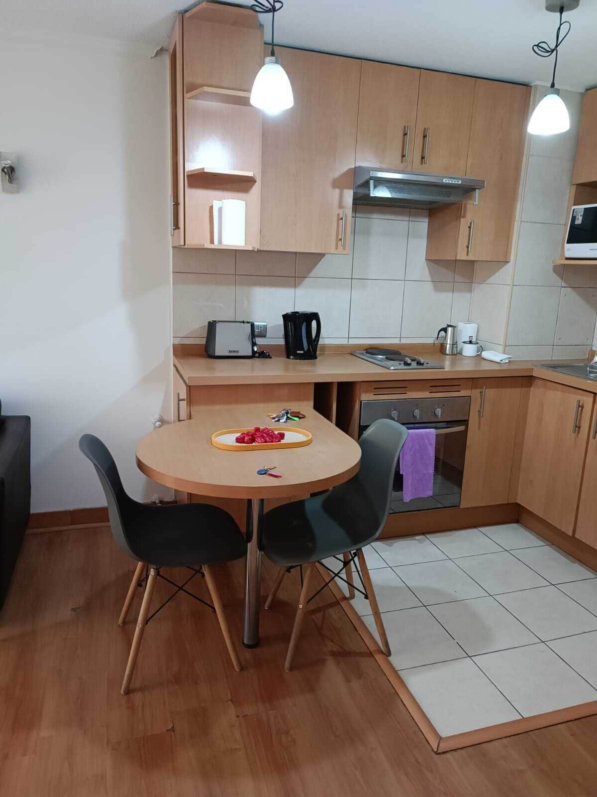 A compact kitchen with wooden cabinets, a round dining table with chairs, a built-in oven, and a microwave.