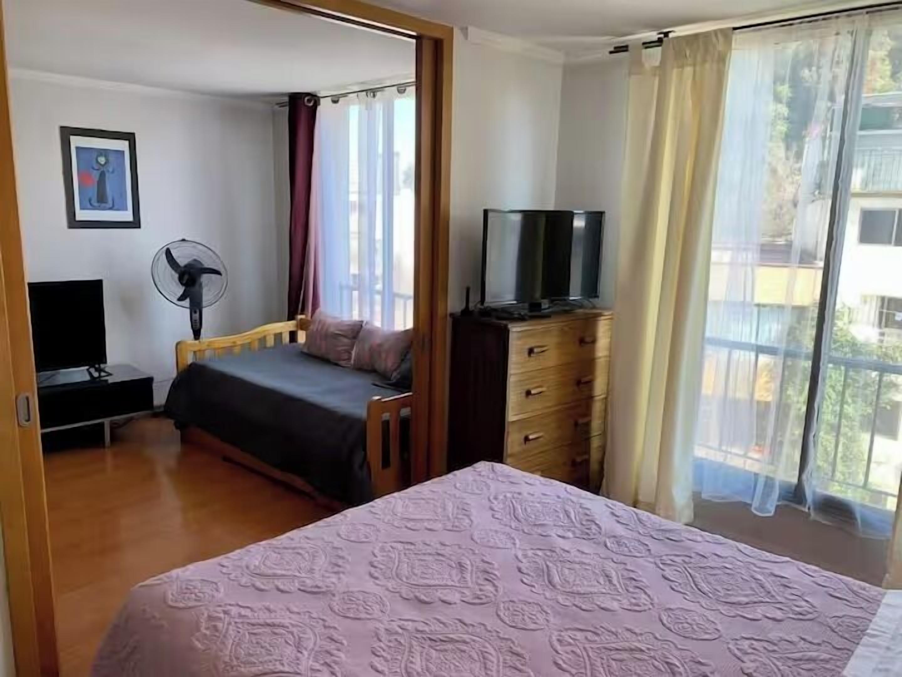 A bedroom with a bed, a dresser, a TV, a fan, and a window with curtains.