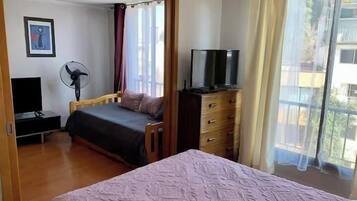 A bedroom with a bed, a dresser, a TV, a fan, and a window with curtains.