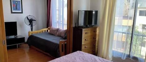 A bedroom with a bed, a dresser, a TV, a fan, and a window with curtains.