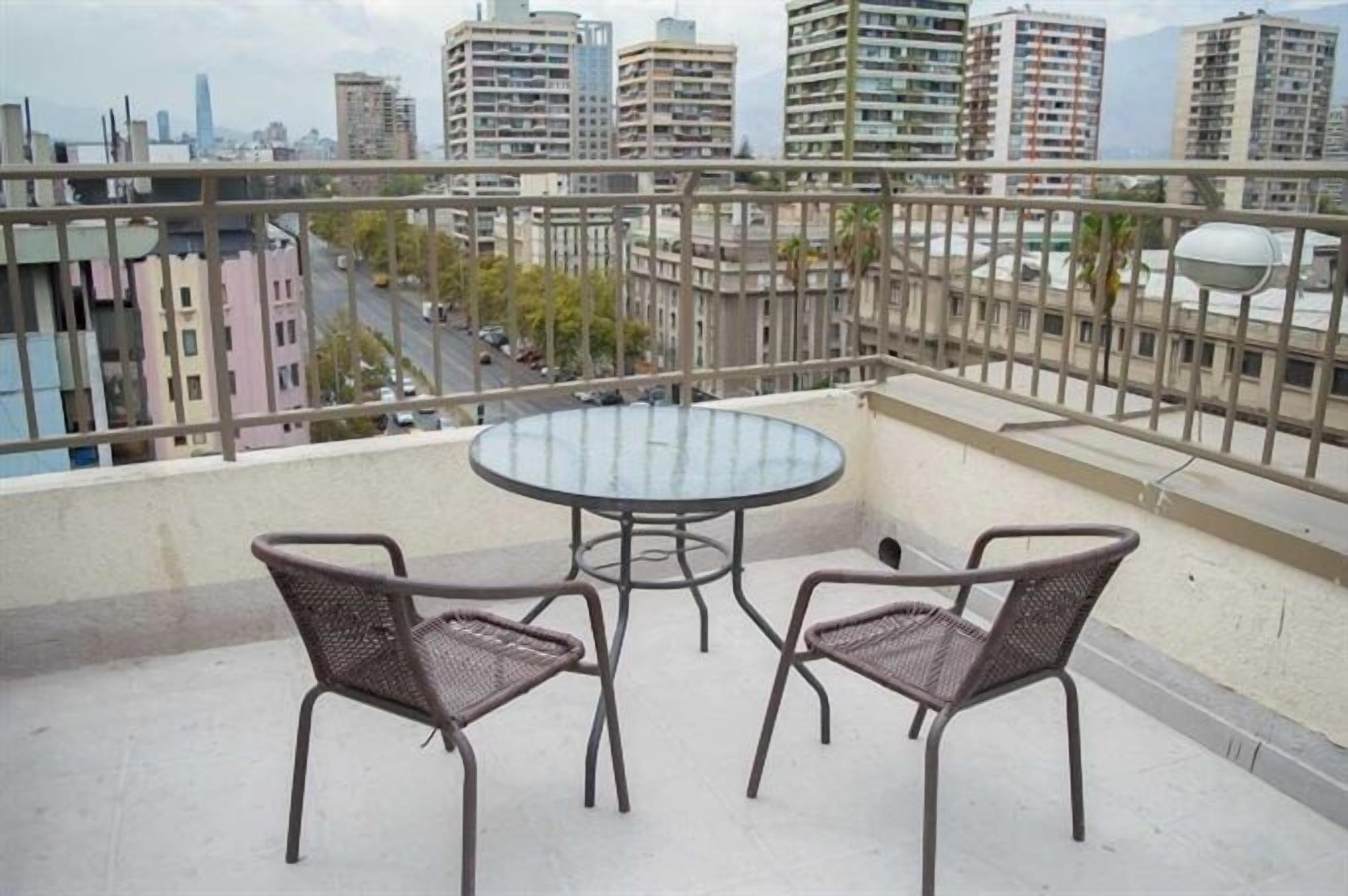 A balcony with a round table and two chairs, overlooking a cityscape with high-rise buildings.