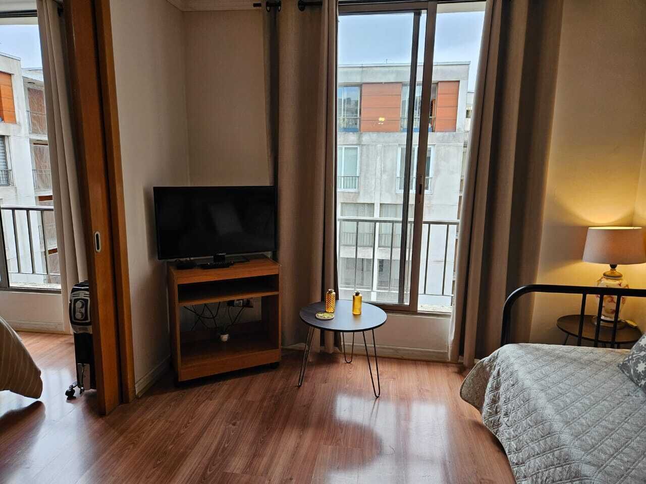 A hotel room with a bed, a TV on a wooden stand, a small round table with a lamp, and a view of buildings through large windows.