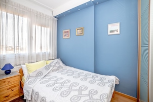 Een slaapkamer met een blauwe wand, een bed met een beddegoed met patroon, een nachtkastje met een lamp en drie ingelijste foto's.