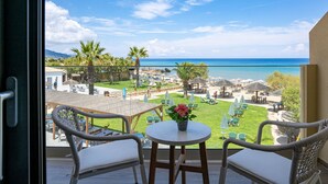 Un balcon avec des fauteuils et une table en osier, donnant sur une plage parsemée de parasols et de chaises longues.
