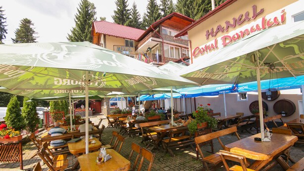 Holztische und -stühle im Freien unter Sonnenschirmen, ein Gebäude mit rotem Dach und ein Schild mit der Aufschrift 'Hotel Donut'.