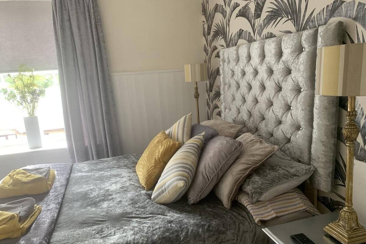 A bed with a tufted headboard, multiple pillows, a bedside lamp, and a remote control on a bedside table.