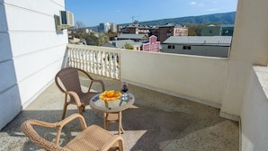A balcony with wicker chairs and a small table, overlooking a residential area with buildings and mountains in the background.