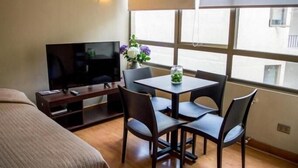 Une chambre d’hôtel avec un lit, un téléviseur posé sur un meuble, une table à manger avec des chaises et une fenêtre donnant sur des immeubles.