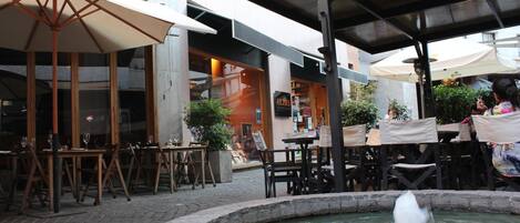 An outdoor dining area with tables and chairs, a fountain, and umbrellas providing shade.