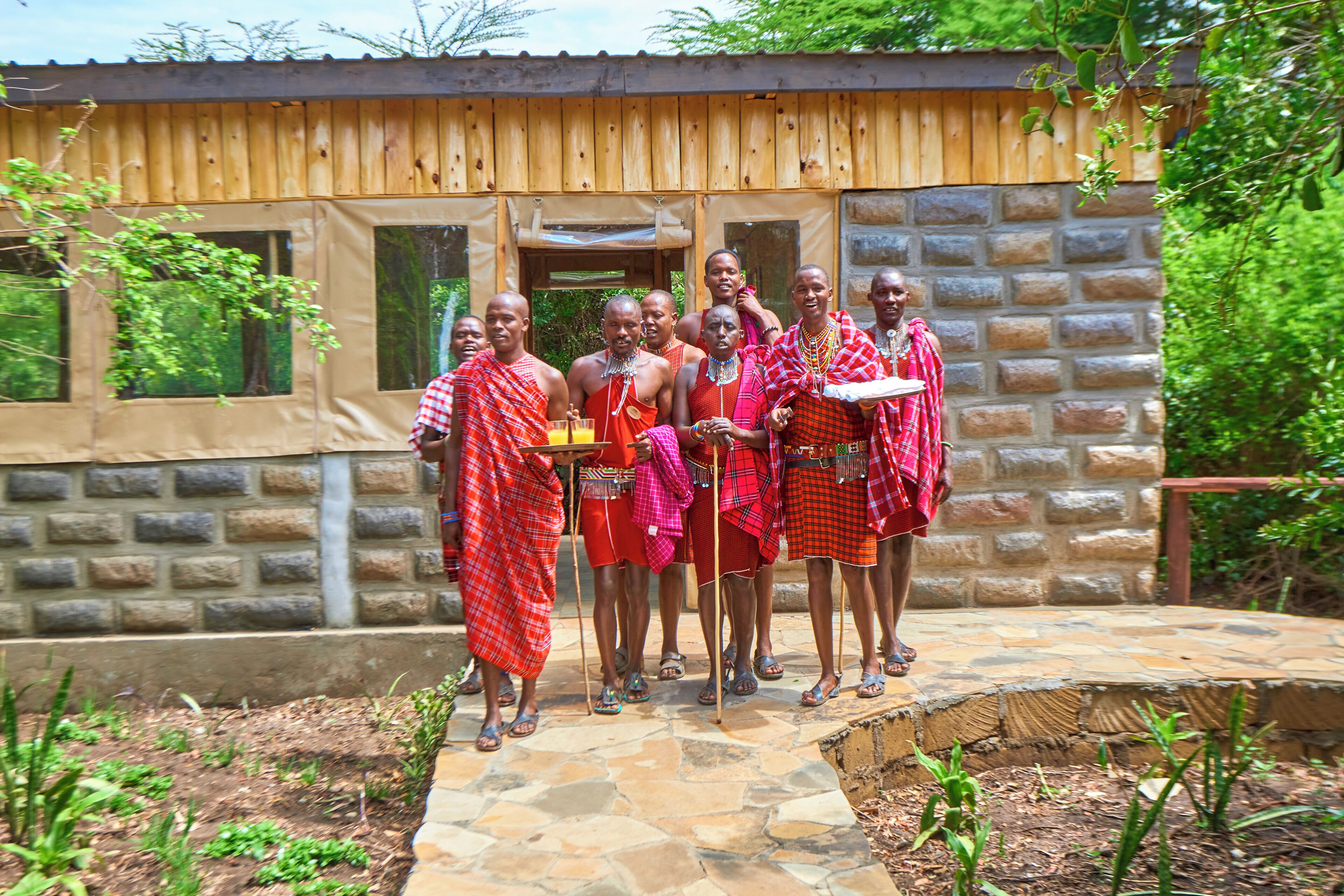 A group of people in traditional attire standing in front of a rustic wooden building with a stone foundation.
