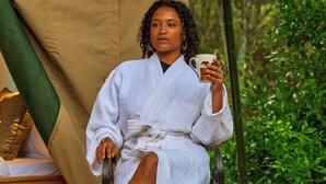 Una persona sentada en una silla en un entorno al aire libre, con una tienda de campaña y vegetación de fondo.