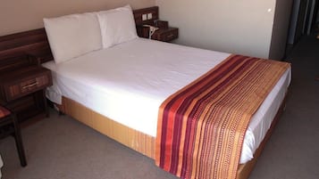 Ein Standard-Hotelzimmer mit einem Einzelbett, einem Nachttisch aus Holz, einer Nachttischlampe und einer Tür rechts.