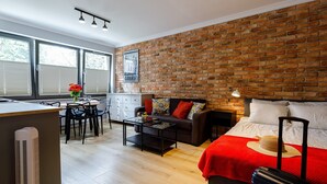 Habitació d'hotel moderna amb paret de maó, llit amb llençol vermell, sofà amb mantell estampat, zona de dinar amb taula i cadires, i cuina amb finestra.