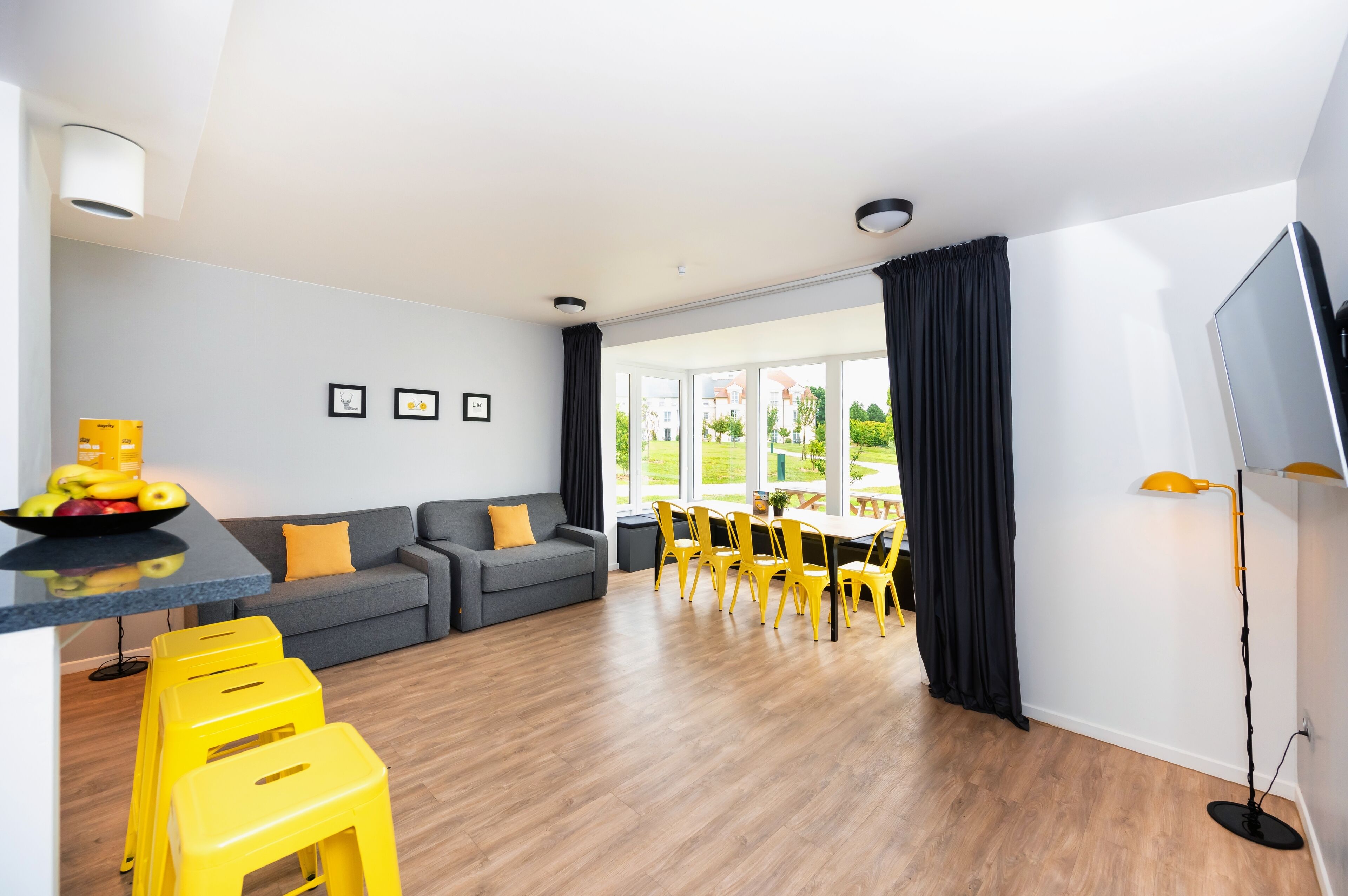 A modern living room with a grey sofa, yellow chairs, a dining table, and a TV.