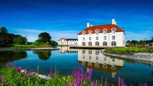 A large white building with a red roof, reflecting in a calm body of water, surrounded by lush greenery and purple flowers. - Staycity Aparthotels, Near Disneyland® Paris (Bailly-Romainvilliers)