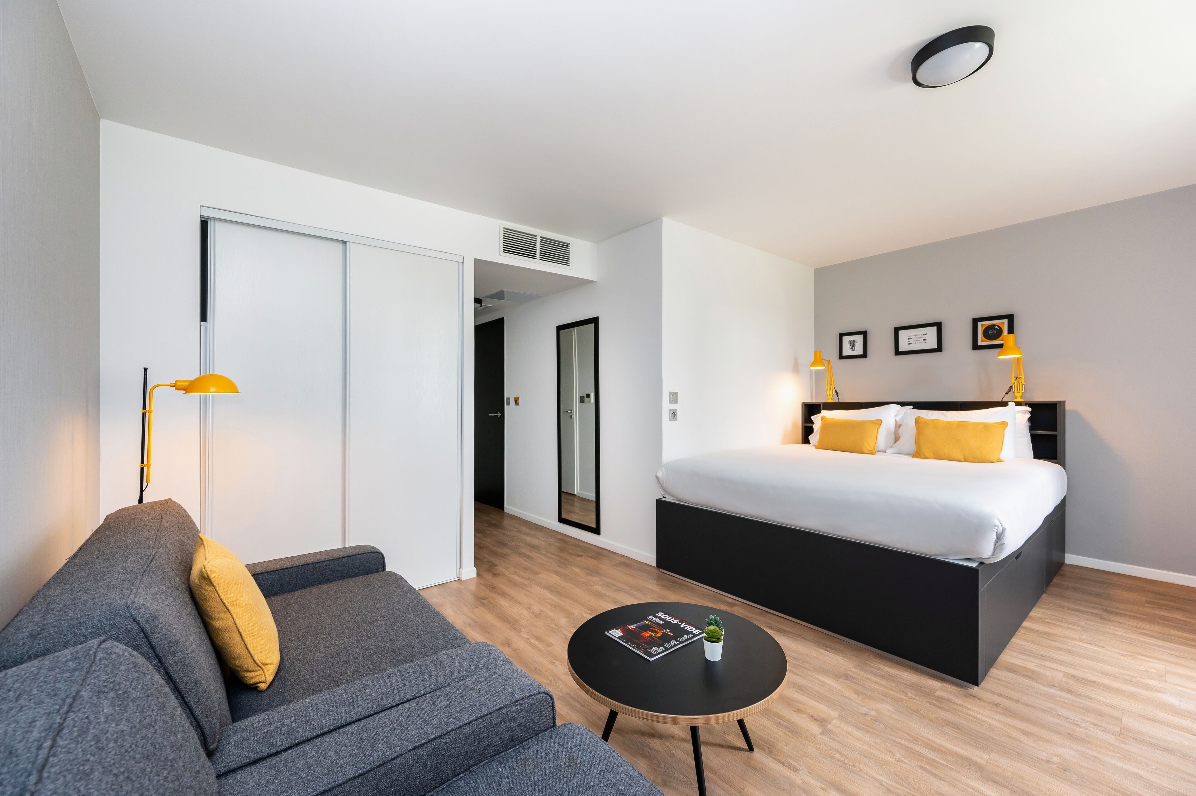 Una camera d'albergo moderna con un divano grigio, un tavolino rotondo con una rivista, un letto con biancheria bianca e cuscini gialli, e un grande specchio sopra il letto.
