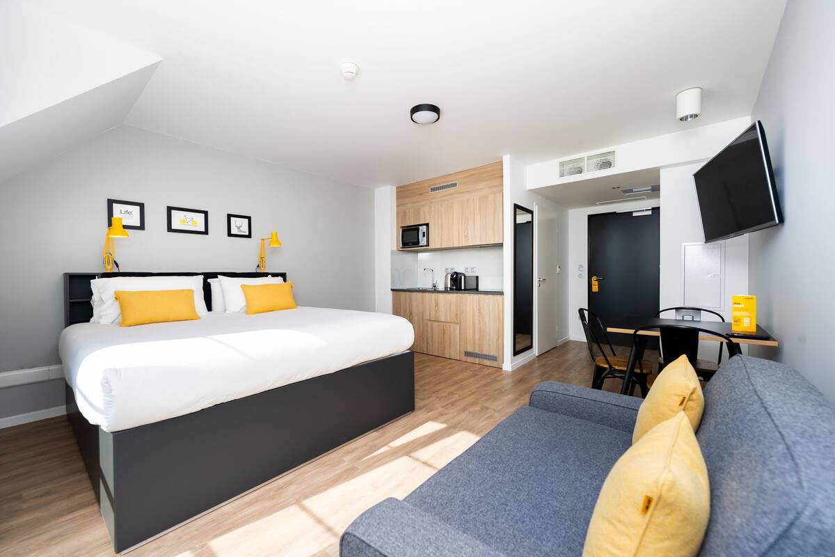 A modern bedroom with a large bed, a sofa, a desk, and a kitchenette.