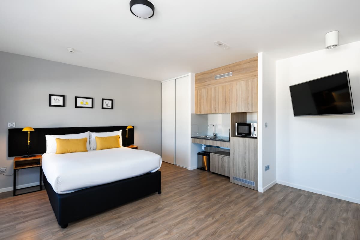 A modern hotel room with a bed, bedside lamps, a flat-screen TV, a small kitchenette, and framed pictures on the wall.
