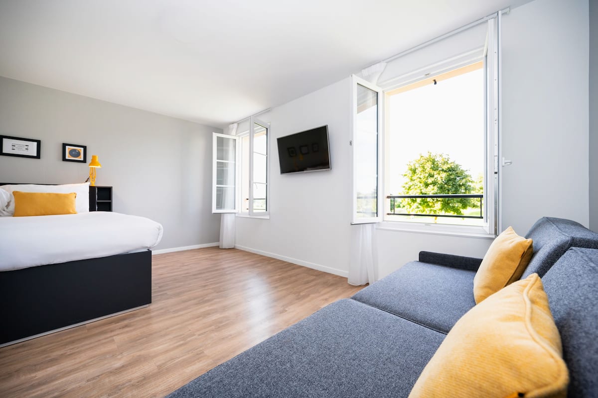 A modern hotel room with a bed, a sofa, a TV, and a balcony with a view of greenery.