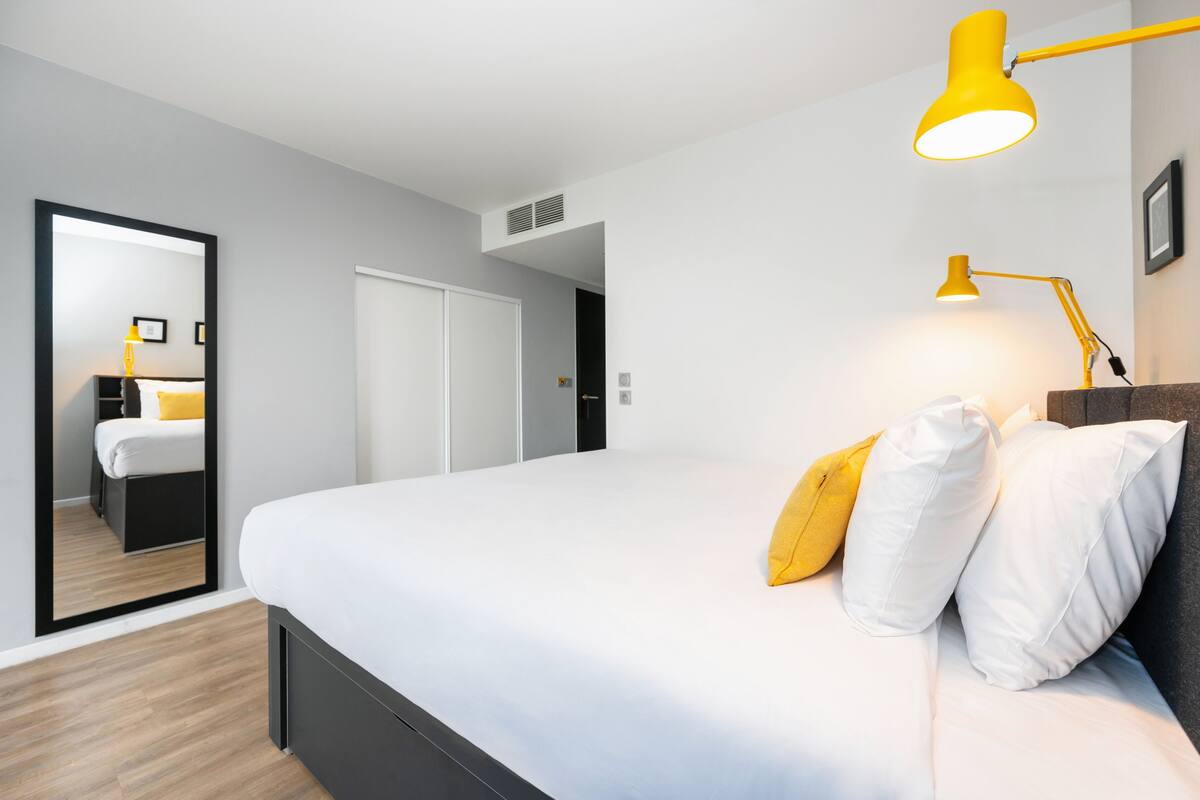 A modern hotel room with a large bed, a mirror, a wardrobe, and a desk with a lamp.